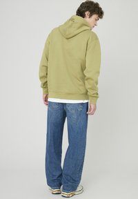 Hoodie vert olive avec un cordon de serrage et un logo dans le dos, associé à un jeans large bleu. Les vêtements présentent une coupe décontractée et une texture douce.