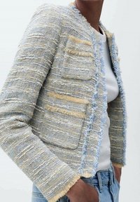Veste en tweed légèrement texturée en bleu pastel et jaune, avec des bords effilochés et deux poches avant. Portée sur un haut blanc.