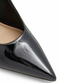 Scarpa nera in vernice con tacco alto, punta appuntita e finitura liscia e lucida. Presenta un interno nero e una soletta beige.