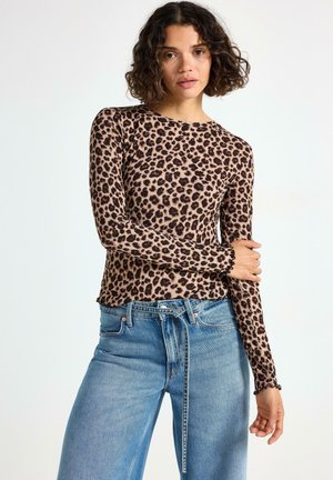 Kvinna med lockigt hår som bär en långärmad topp med leopardtryck och ljusblå högmidjade jeans, poserar med ena armen i kors.