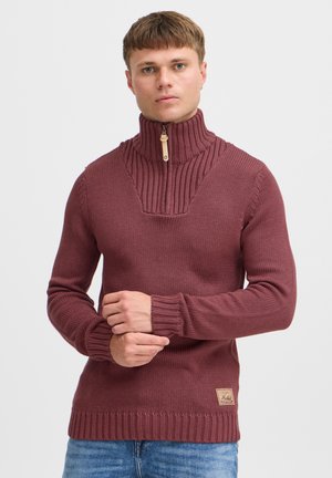 Bordo pletena dukserica s visokim ovratnikom i polu-zip suknjom, s rebrastim detaljima i tkanom etiketom na donjem rubu.