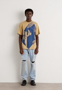 Beige t-skjorte med blå grafisk design som viser en fugl og abstrakte former, kombinert med lyseblå slitte jeans og svarte sko.