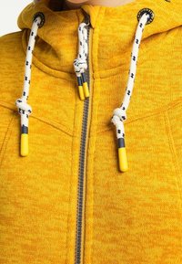 Žlutá mikina na zip vyrobená z texturované tkaniny. Má stahovací šňůrku s žlutými a modrými akcenty a malé logo poblíž límečku.