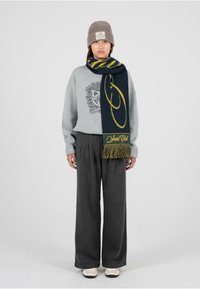 Szara dziana czapka, oversize szarysweatshirt z logo, ciemnoszare spodnie szerokie w nogawkach oraz granatowy szalik z żółtym wzorem i frędzlami.