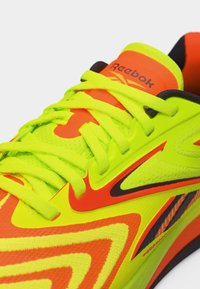 Κοντινό πλάνο σε αθλητικό παπούτσι Reebok σε νεόν κίτρινο και πορτοκαλί χρώμα, με κορδόνια ίδιου χρώματος και μαύρες λεπτομέρειες, σε λευκό φόντο.
