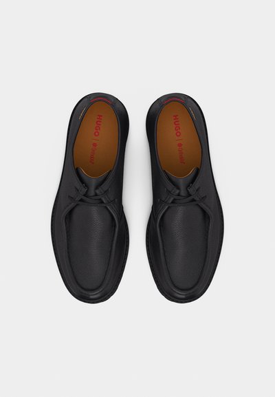 Paire de chaussures habillées noires en cuir à lacets avec semelles intérieures marron marquées "HUGO" sur un fond clair uni.
