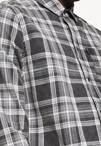 Camicia a quadri grigio e nera, con colletto, chiusura con bottoni e taschino sul petto. Il tessuto ha una consistenza morbida con motivi distinti.