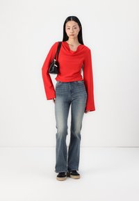 Rød genser med hetteløsning og lange ermer, kombinert med lyseblå utsvingte jeans og svarte sneakers, mens hun holder en liten svart håndveske. Hvit bakgrunn.