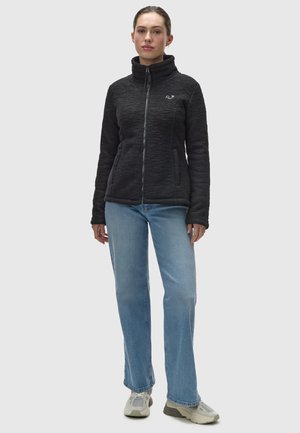 Schwarze Fleecejacke mit hohem Kragen, Frontreißverschluss und zwei Seitentaschen. Modell trägt hellblaue gerade Jeans und Sneakers.