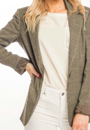 Femme aux longs cheveux blonds portant un chemisier beige, un pantalon blanc et un blazer marron à carreaux, une main sur la hanche.
