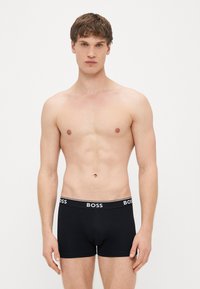 Cuecas boxer pretas com cós de logótipo, design ajustado e tecido suave. Modelo está em pé, sem camisa, contra um fundo branco liso.