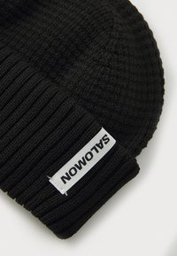 Zwarte gebreide beanie met een geribbelde textuur en een wit label met "SALOMON" in zwarte letters.
