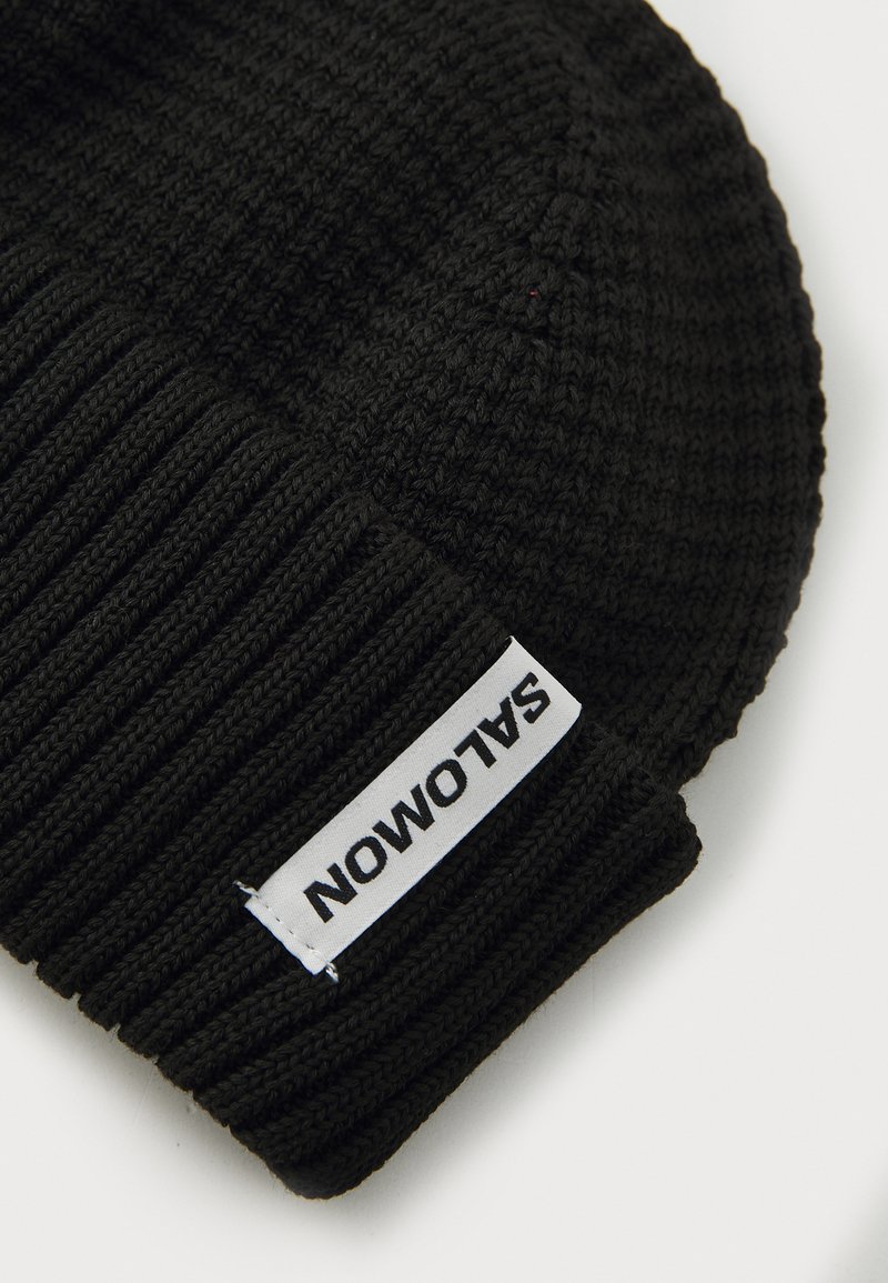 Berretto di maglia nera con texture a costine e un'etichetta bianca con la scritta "SALOMON" in font nero.