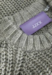 Pull gris clair en tricot avec col côtelé et une étiquette violette marquée "JJXX" près de l'encolure.