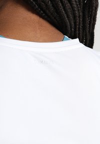 Gros plan sur le cou et le haut du dos d'une personne aux cheveux foncés et tressés, portant une chemise blanche avec un texte discret « CLIMALITE » près du col.