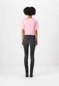 Růžové bavlněné tričko s krátkými rukávy, které je zkombinováno s tmavě šedými skinny džínami. Na nohou jsou černé platformové kotníkové boty. Pohled zezadu na jednobarevném pozadí.