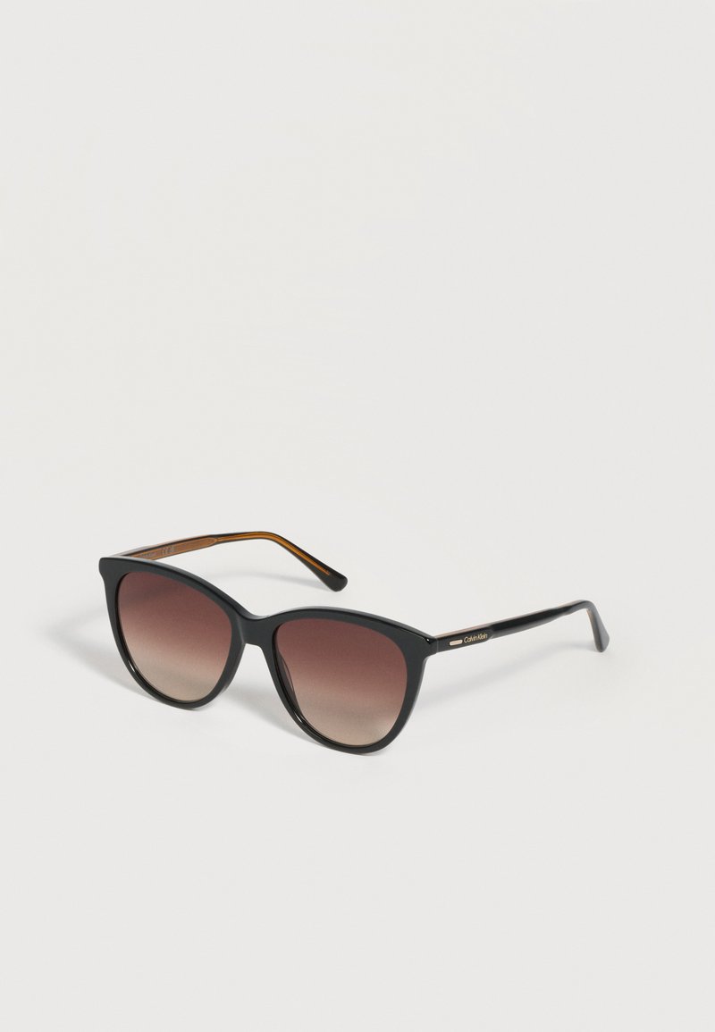 Lunettes de soleil noires forme œil de chat avec verres dégradés marron et branches fines, logo Calvin Klein sur les branches.