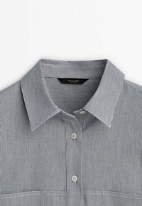 Męska koszula zapinana na guziki w wąskie czarne i białe pionowe paski z kołnierzykiem w kształcie punktu, ukazująca górną przednią część na białym tle.