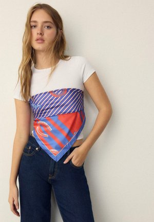 Jeune femme aux longs cheveux blonds portant un t-shirt blanc, un jean bleu et une écharpe rouge et bleue à motifs nouée autour de sa poitrine.