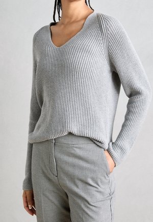 Femme portant un pull en maille côtelée gris clair avec un col en V et un pantalon à carreaux gris, une main dans la poche, posant sur un fond uni.