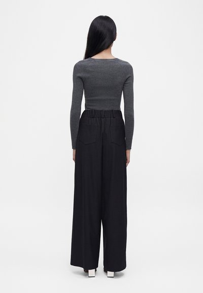 Personne aux longs cheveux noirs, vue de dos, portant un haut gris ajusté à manches longues et un pantalon noir ample taille haute avec des poches arrière.