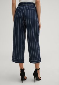 Pantaloni cropped blu navy con righe verticali bianche, vita elastica e tasca posteriore. Il tessuto è liscio e con un taglio sartoriale. Indossati con tacchi neri.