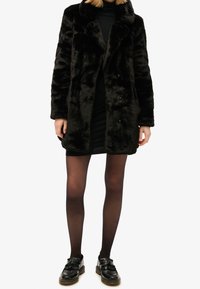 Frau in einem schwarzen Kunstpelzmantel, schwarzen durchsichtigen Strumpfhosen, schwarzem Mini-Kleid und schwarzen Lederloafers vor einem weißen Hintergrund.