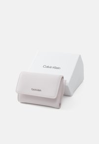 Calvin Klein vaaleanpunainen nahkalompakko, jossa on läppäsuljenta, hopeinen logoelementti ja kompakti suorakulmainen muoto, valkoisen laatikon vieressä.