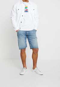Vit denimjacka över en vit t-shirt med en färgglad grafik, kombinerad med ljusblå denimshorts och vita sneakers.
