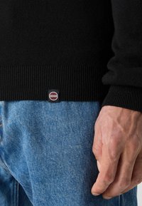 Μαύρο πλεκτό πουλόβερ με ριμπ μανσέτες και στρίφωμα, συνδυασμένο με ανοιχτό μπλε τζιν. Ορατό λογότυπο κοντά στο στρίφωμα. Απαλή υφή.