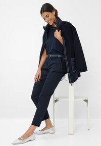 Cappotto imbottito blu navy sopra una maglia blu navy, abbinato a pantaloni blu navy e ballerine argento chiaro. Texture lisce e design minimalista.