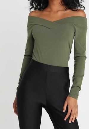 Femme portant un haut kaki à épaules dénudées et manches longues, associé à un pantalon noir ajusté taille haute, debout avec une main détendue.