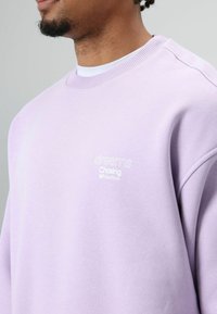 Sweat-shirt lavande en tissu doux avec un col côtelé et un texte brodé en blanc : "dreams Chasing NOT deadlines."