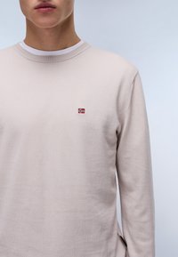 Lichtbeige trui met een ronde halslijn, lange mouwen en een kleine geborduurde Noorse vlag op de borst. Zachte textuur, minimalistisch ontwerp.