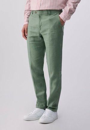 Uomo con pantaloni verde chiaro a gamba dritta, camicia rosa a righe infilata e sneakers bianche, in piedi con una mano nella tasca.