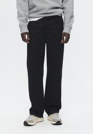 Persona che indossa pantaloni neri a gamba dritta, una felpa grigia, una maglietta bianca intima e scarpe da ginnastica beige, in piedi davanti a uno sfondo semplice.