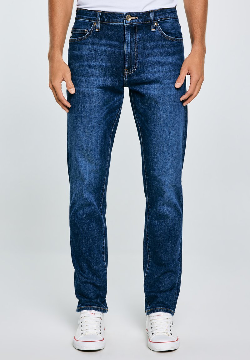 Jeans de mezclilla de color azul oscuro con corte recto, que cuentan con cremallera, cinco bolsillos y detalles de costura sutiles. Combinados con zapatillas blancas.