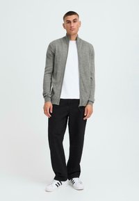 Grå zip-genser med ribbestrikkede mansjetter, brukt over en hvit t-skjorte, kombinert med svarte bukser og hvite joggesko med svarte striper.