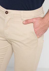 Pantaloni bej cu închidere prin nasture și bucle pentru curea, mână parțial în buzunarul din față, purtați cu o cămașă de culoare bleumarin înfiptă.