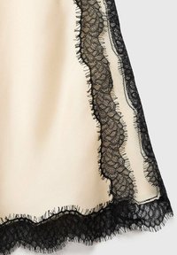 Tissu beige avec des bordures en dentelle noire, présentant des motifs floraux complexes et des bords à franges le long de l'ourlet et des côtés pour ajouter de la texture.
