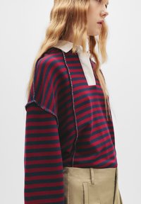 Gestreifter Pullover in Rot und Navy mit einem weißen Kragen, komfortabler Schnitt und sichtbaren Nähten an den Kanten. Kombiniert mit beigen trousers.