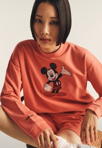 Giovane con capelli scuri corti che indossa una felpa corallo e un set di pantaloni corti con una grande grafica di Topolino allegra sul davanti.