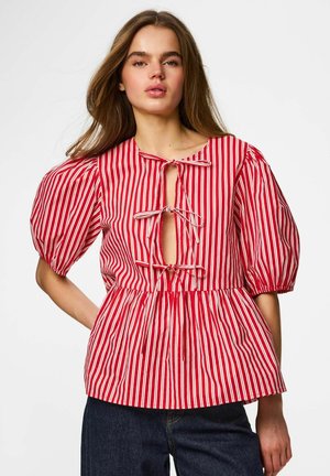 Kobieta nosząca czerwoną i białą bluzkę w pionowe paski z bufkami i trzema związanymi kokardami z przodu, zestawiona z ciemnymi dżinsami.