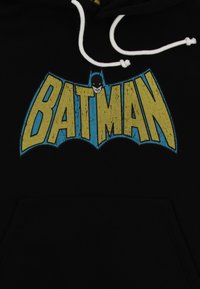 Felpa nera con cappuccio caratterizzata da un grande logo giallo "BATMAN" in stile rovinato con un accento blu e un volto stilizzato di Batman sopra il testo. Cordini bianchi.