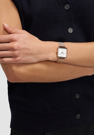 Goudkleurig rechthoekig horloge met een wit wijzerplaat, zwarte uurmarkeringen en een metalen mesh band, gedragen om een pols met een donkere gebreide cardigan.