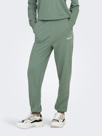 Pantalon de survêtement vert en tissu doux avec poches latérales, arborant un logo "LIMITLESS" imprimé ; porté avec des baskets blanches et noires épaisses.