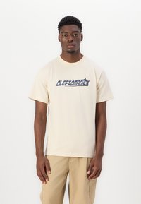 Beige cotton t-shirt with short sleeves, featuring a blue graphic logo "CLEPTOMANIX" across the chest. Simple design, relaxed fit.