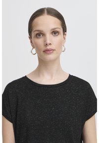 Heledate nahaga ja siniste silmadega naine, kes kannab musta täpilist lühikeste varrukatega särki ja hõbedasi rõngakujulisi kõrvarõngaid, seisab näoga otse ja tal on neutralt väljendatud nägu.