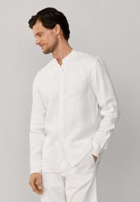 Hombre con camisa blanca de lino con cuello mandarín y pantalones blancos a juego, de pie con una mano en el bolsillo, mirando hacia abajo y sonriendo ligeramente.