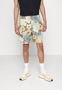 Spodenki w kwiatowy wzór w ziemistych odcieniach brązu, niebieskiego, zielonego i kremowego, z elastycznym pasem. Zestawione z białymi trampkami i skarpetkami.